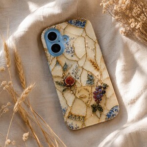 A mosaic phone case adorned with figs, grapes, and wheat sits on rumpled cream fabric next to dried wheat stems.