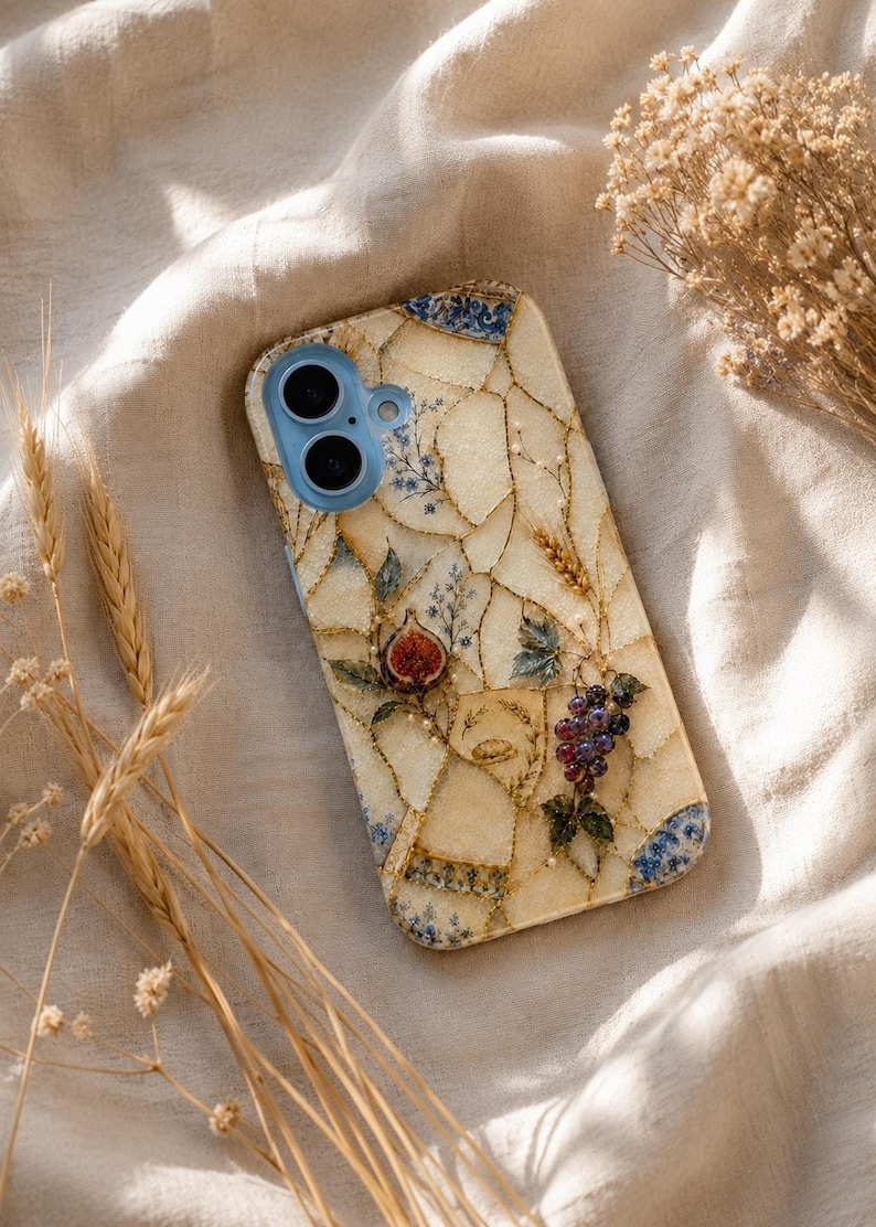 A mosaic phone case adorned with figs, grapes, and wheat sits on rumpled cream fabric next to dried wheat stems.