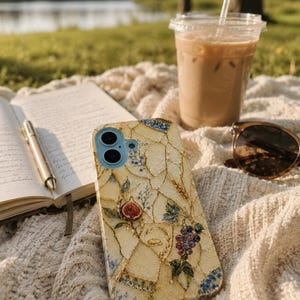 A mosaic phone case lies on a textured blanket near a notebook, pen, iced coffee, and sunglasses outdoors.
