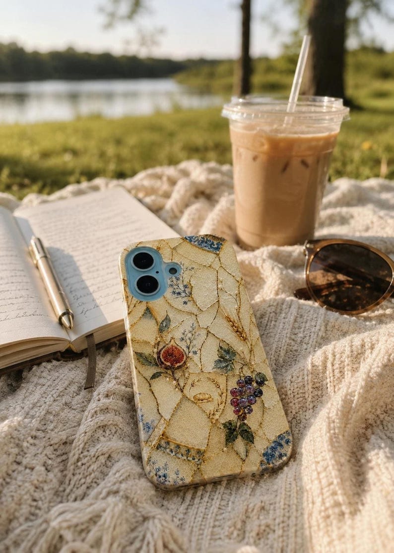 A mosaic phone case lies on a textured blanket near a notebook, pen, iced coffee, and sunglasses outdoors.