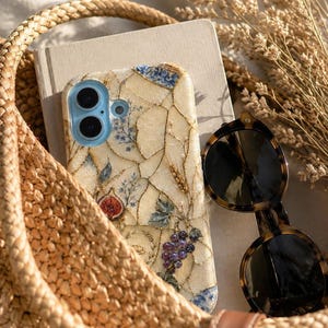 A woven straw bag holds a notebook, sunglasses, and a mosaic phone case decorated with fruit designs.