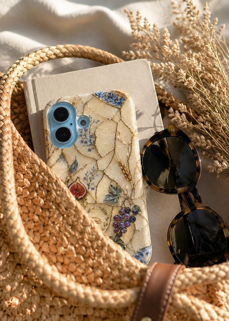 A woven straw bag holds a notebook, sunglasses, and a mosaic phone case decorated with fruit designs.