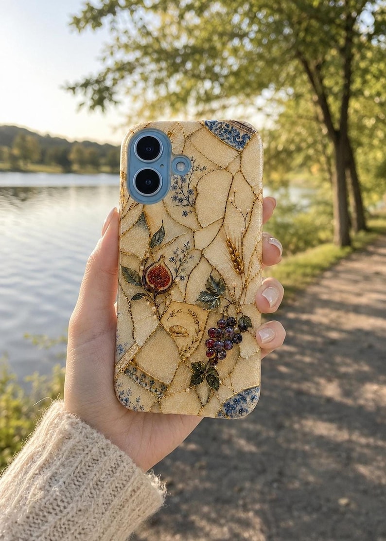 A hand holds up a mosaic-patterned phone case featuring figs, grapes, and wheat against a scenic outdoor path and water backdrop.