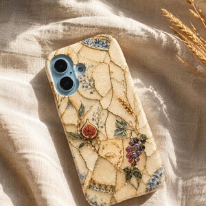 A mosaic-style phone case featuring figs and grapes rests on a textured cream fabric surface beside dry wheat stalks.