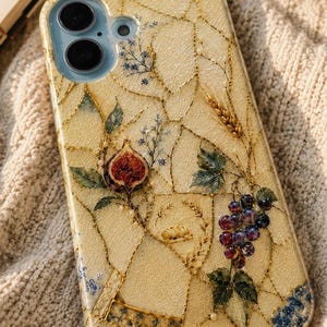 Close-up of a mosaic phone case featuring detailed paintings of figs, purple grapes, and wheat on a cream knit background.