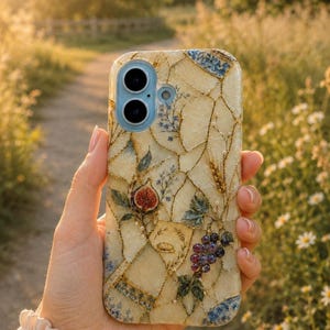 A hand holds a mosaic-patterned phone case outdoors along a dirt path flanked by greenery and small white flowers in warm sunlight.