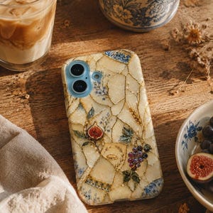 A phone case with a cream-colored kintsugi mosaic pattern featuring botanical motifs like grapes, figs, and wheat rests on a wooden table beside a glass of iced coffee and a blue floral mug.