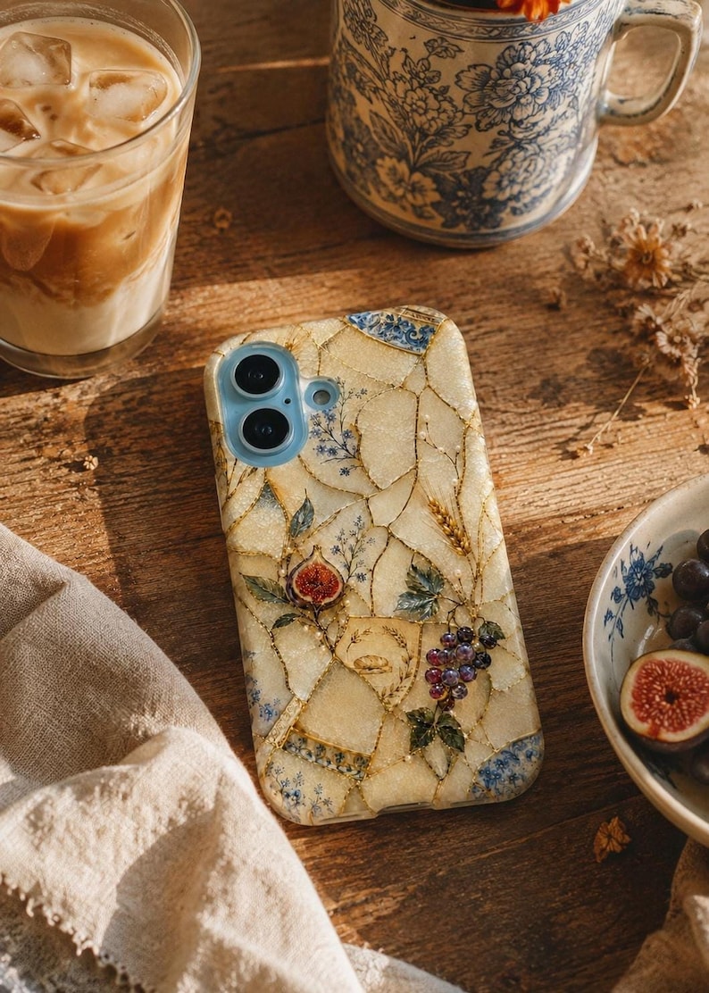A phone case with a cream-colored kintsugi mosaic pattern featuring botanical motifs like grapes, figs, and wheat rests on a wooden table beside a glass of iced coffee and a blue floral mug.
