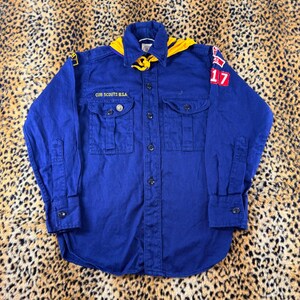 May include: A long-sleeved blue Cub Scouts B.S.A. button-down shirt featuring a yellow neckerchief, two front chest pockets, and various embroidered patches on the sleeves, including the number 17 in red and white on the left shoulder.