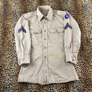 May include: A long-sleeved tan military-style button-down shirt featuring two chest pockets, shoulder epaulets, and chevron patches on both sleeves. The left shoulder displays a circular patch with a blue border and a red letter A in the center.