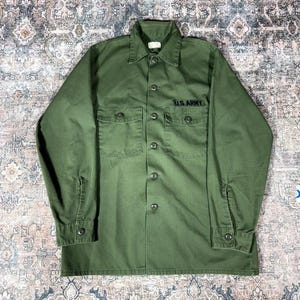 Può includere: Una camicia a maniche lunghe color verde oliva con abbottonatura frontale, colletto a punta, due tasche sul petto con patta e polsini abbottonati. La scritta U.S. ARMY è ricamata in filo nero sopra la tasca destra sul davanti.