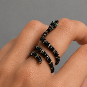 May include: A coiled snake ring featuring black beads arranged in a textured pattern with thin gold-colored bands interspersed throughout the body. The snake head is detailed with two small, vibrant green circular eyes, creating a distinct contrast.