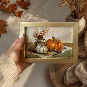 May include: An oil painting in a gold frame depicting a still life scene with a large orange pumpkin, a smaller white pumpkin, and a dark vase filled with orange berries, all resting on a light-coloured surface with scattered autumn leaves.