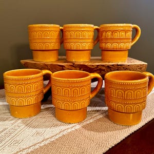 Può includere: Un set di sei tazze in ceramica color giallo senape abbinate. Ogni tazza presenta un motivo in rilievo con archi ripetuti e linee tratteggiate sulla parte superiore, una base cilindrica liscia e un manico curvo sul lato destro.