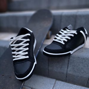 May include: A pair of black low-top sneakers with white laces and a white midsole. The shoes feature a smooth black leather-like toe box and side panels with black suede-like overlays. The sneakers are positioned near a dark skateboard.