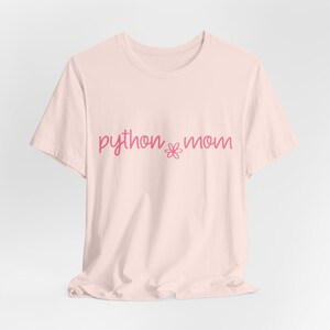 May include: A light pink short-sleeved t-shirt featuring the text "python mom" in a dark pink cursive script. Between the two words is a small, simple line-drawn flower icon. The shirt has a crew neckline and a standard, relaxed fit.