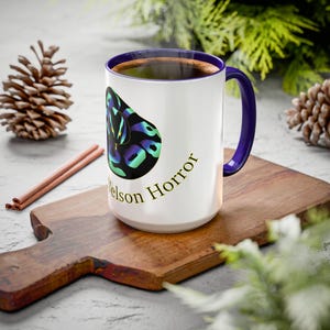 May include: A white ceramic mug with a dark purple interior and handle. The exterior features a graphic of a black snake with iridescent blue and green scales, accompanied by the text "Nelson Horror" printed in a gold-colored serif font.