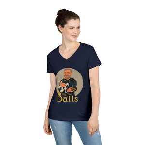 May include: A navy blue V-neck t-shirt featuring a circular graphic of a person holding a black and white cat. Below the graphic, the text reads "Hug Your Balls" in a gold-colored font. The shirt has short sleeves and a standard fit.