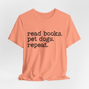May include: A peach-colored short-sleeve t-shirt featuring the black text "read books. pet dogs. repeat." printed in a simple, sans-serif font across the center of the chest. The garment has a classic crew neckline and a standard straight hem.