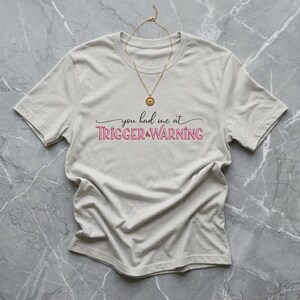 May include: A light beige short-sleeved t-shirt featuring the black and pink text you had me at TRIGGER WARNING printed on the chest. A thin gold-toned chain necklace with a small circular pendant rests on the neckline of the shirt.