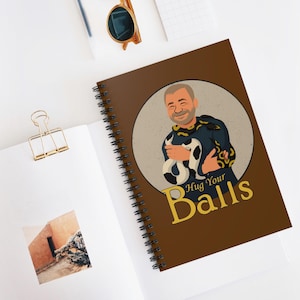 May include: A brown spiral-bound notebook featuring a circular illustration of a smiling person holding a black and white patterned snake. The text "Hug Your Balls" is printed in large yellow letters below the circular graphic design.
