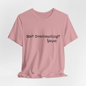May include: A dusty rose short-sleeved t-shirt featuring black text printed on the front. The text reads, "Me? Overreacting? Never" in a combination of a standard serif font for the first two phrases and a handwritten script font for the word Never.