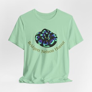 May include: A light mint green short-sleeve t-shirt featuring a graphic of a coiled snake with iridescent blue and purple scales. The text Bridgett Nelson Horror is printed in a curved gold font directly underneath the snake illustration.