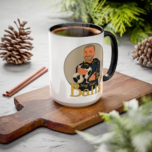 May include: A white ceramic mug with a black interior and handle, featuring a circular graphic of a person holding a black and white snake. Below the graphic, the text reads "Hug Your Balls" in gold lettering, with a dark rim and handle.