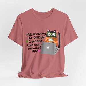 May include: A dusty rose t-shirt featuring a graphic of a black cat wearing green glasses and an orange striped sweater, sitting at a laptop with a drink. The text reads: ME tracking the ORDER I placed two damn minutes ago, with small hearts.