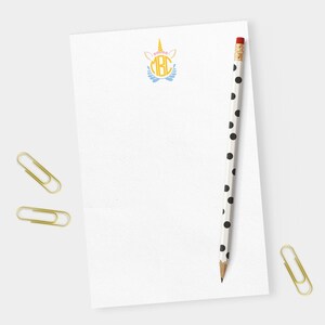 May include: A white notepad with a unicorn and the letters "MBC" in a circle. A black and white polka dot pencil is on the notepad.