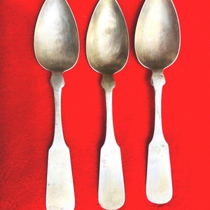 May include: Three antique silver spoons with oval bowls and elongated handles are arranged vertically against a vibrant red background. The spoons have a classic design, reflecting a vintage aesthetic. The spoons are likely for serving or eating.