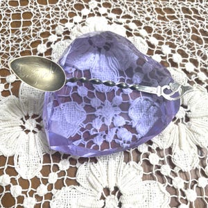 May include: A silver spoon with a twisted handle rests on a purple, heart-shaped object. The spoon's bowl has etched details. The objects are displayed on a white lace doily with floral patterns. The background is a wooden surface.