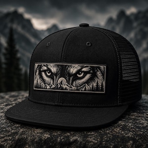 May include: A black trucker hat featuring a rectangular patch on the front. The patch displays a detailed illustration of a wolf's eyes peering over a mountain range with a full moon and pine trees in a monochromatic black and white color scheme.
