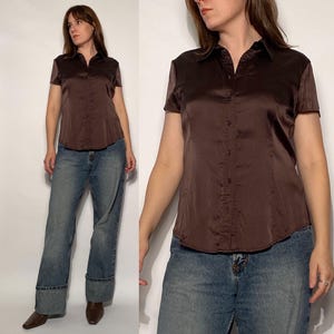 May include: A brown satin short-sleeved shirt with a collared neckline and button-down front. The shirt is worn with blue denim jeans with a wide leg and a folded cuff. The model is wearing brown shoes. The shirt is a classic style.