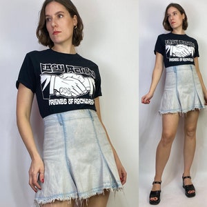 May include: A woman is wearing a black t-shirt with the text "Easy Action Friends of Rockers" in white. She is also wearing a light blue denim skirt with a frayed hem.