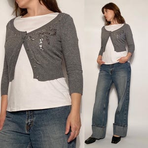 May include: A grey, cropped cardigan with a sequined bow design. The cardigan is buttoned and worn over a white scoop-neck top. The model is wearing blue jeans.