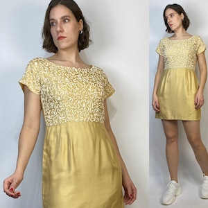 May include: A yellow short-sleeved dress with white embroidered detailing on the bodice. The dress has a scoop neckline and a straight skirt.