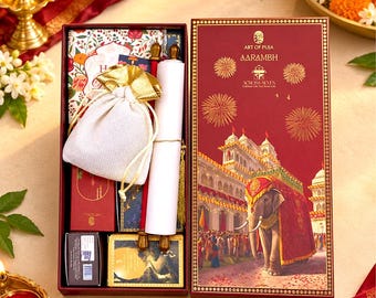 May include: A rectangular gift box in deep red featuring a gold illustration of an elephant and a palace. Inside are a white drawstring pouch, a rolled scroll with gold finials, a small red box, and a card with the text Art of Puja Aarambh.