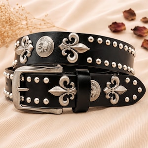 Può includere: Una cintura in pelle nera con finiture in metallo argentato, tra cui una fibbia rettangolare, file di piccoli borchie rotonde, medaglioni circolari in rilievo raffiguranti un teschio con elmo e decorazioni metalliche a forma di fleur-de-lis.