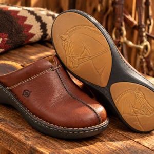 May include: A pair of brown leather slip-on shoes with white contrast stitching. One shoe is shown from the side, while the other is positioned to display the tan rubber sole, which features an embossed horse head design and black accents.