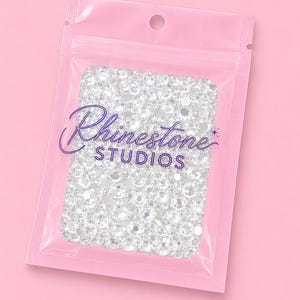 May include: A clear plastic resealable pouch containing numerous small, round, faceted clear rhinestones. The front of the bag features the purple text Rhinestone STUDIOS printed in a stylised font with a small star icon above the letter S.