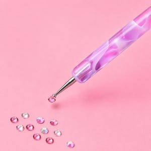 Op de afbeelding: Een diamond painting pen met een paars en wit gemarmerde acryl handgreep en een zilverkleurige metalen punt. De punt houdt een kleine roze strass-steen vast, terwijl er diverse andere roze en doorzichtige strass-steentjes verspreid liggen.