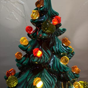 May include: A dark green ceramic Christmas tree featuring a glossy finish and molded branch details. The tree is decorated with numerous translucent, acorn-shaped light bulbs in shades of amber, orange, red, and yellow that glow from within.