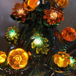 May include: A dark green ceramic tree sculpture featuring illuminated translucent orange and yellow accents shaped like pumpkins and autumn leaves. The decorative piece glows from within, highlighting the glossy finish and the detailed textures of the foliage and gourds attached to the branches.