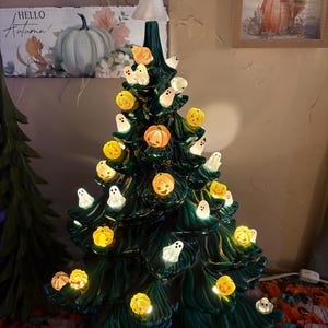 May include: A dark green ceramic tabletop tree decorated with illuminated miniature orange pumpkins and white ghosts. The tree is topped with a white ghost figure wearing a small red bow. The lights are embedded throughout the branches.