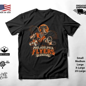 Könnte beinhalten: Ein schwarzes Kurzarm-T-Shirt mit einer Grafik eines Eishockey-Torwarts in orange-schwarzer Ausrüstung mit Flügeln, einem Puck und der Liberty Bell vor einem Mond. Der Text lautet Philadelphia Flyers und Fly by Fright.
