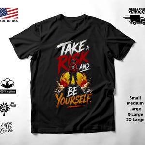 May include: A black short-sleeved t-shirt featuring a graphic print of a figure standing on a mountain peak against a large orange sun. The text reads "TAKE A RISK AND BE YOURSELF" in white, red, and yellow stylized lettering across the chest.