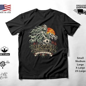 May include: A black short-sleeve t-shirt featuring a graphic print of a woman with flowing hair holding a soccer ball in front of mountains and a sun. The text reads Denver Summit FC and Summit of Spirits, with floral accents below the text.