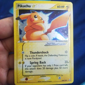 May include: A yellow Pikachu trading card with 60 HP, featuring an illustration of Pikachu holding a sword. The card text includes Thundershock and Spring Back attacks, along with the rule: You can't have more than 1 Pokémon star in your deck.