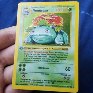 Peut inclure: Une carte à collectionner rectangulaire représentant le personnage vert Florizarre avec une grande fleur rouge sur le dos. La carte affiche 100 PV, le texte Pokémon Power: Energy Trans, Lance-Soleil 60 et un tampon 1st Edition sur bordure jaune.
